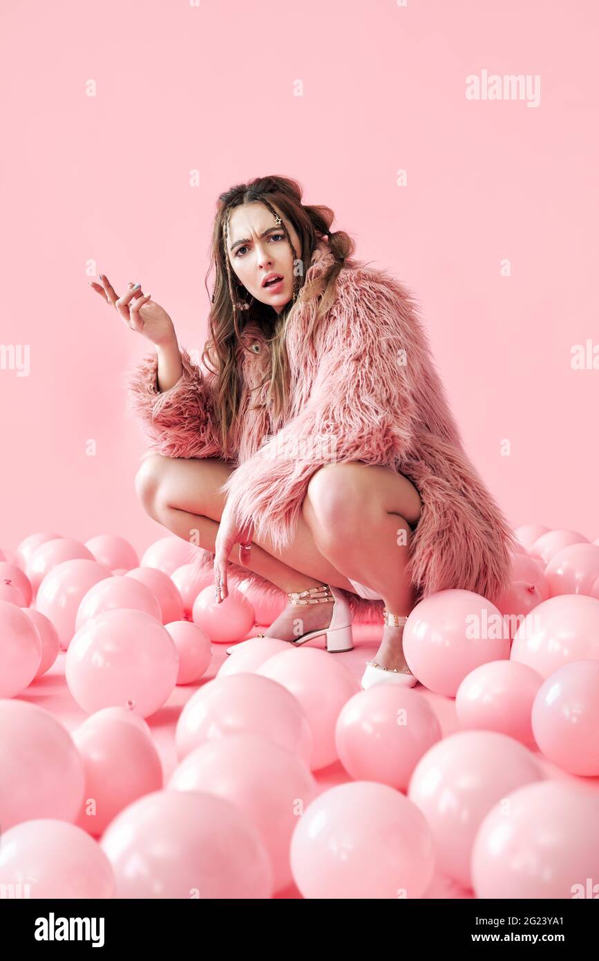 Displeased hipster woman sitting in many pink balloons background. Emotion concept Stock Photo