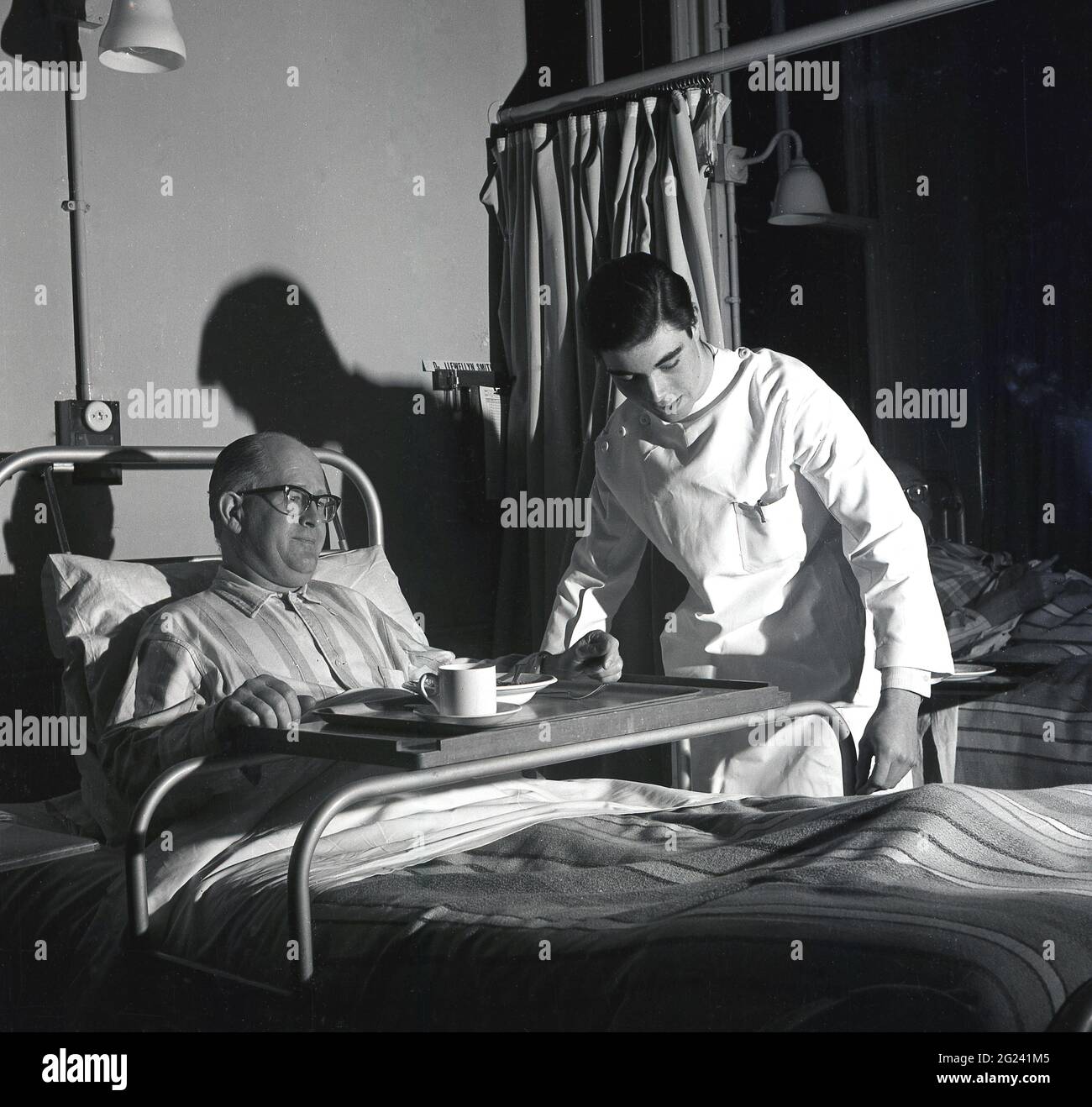 1960s, historical, young male doctor working an evening a ward shift helping make an elderly male patient more comfortable in his bed, by adjusting the  frame of the metal bed holder for his dinner tray, Scotland, UK. Stock Photo