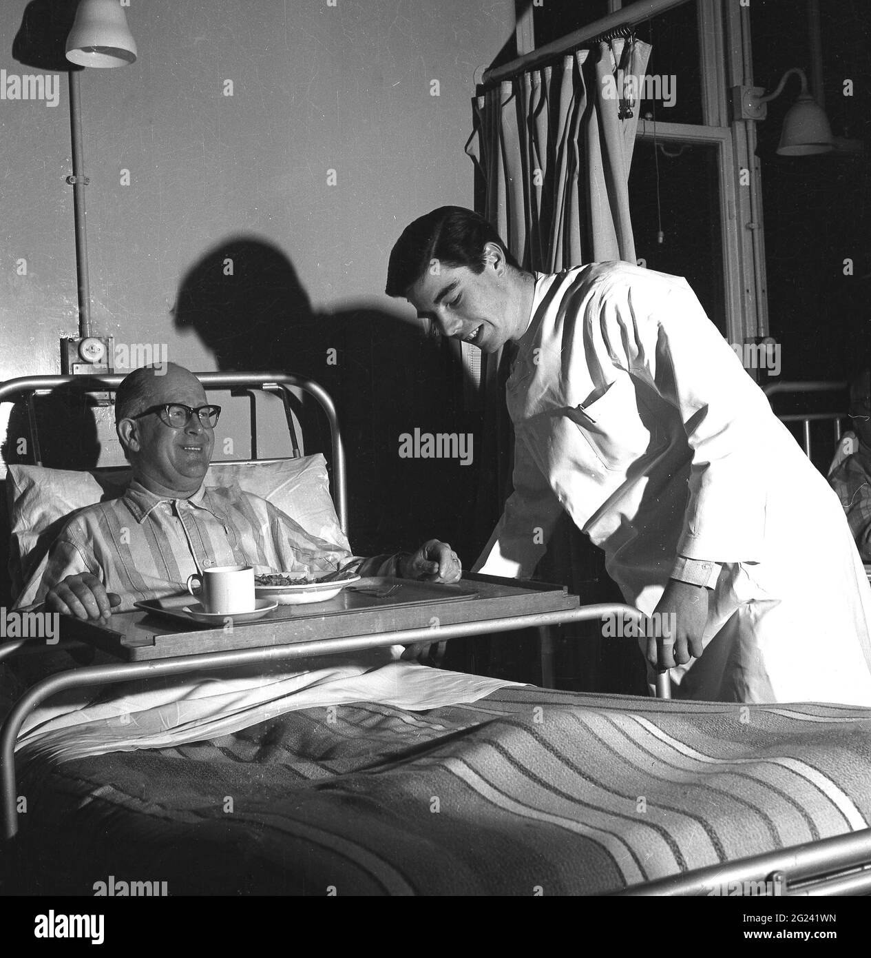 1960s, historical, young male doctor working an evening a ward shift helping make an elderly male patient more comfortable in his bed, by adjusting the  frame of the metal bed holder for his dinner tray, Scotland, UK. Stock Photo