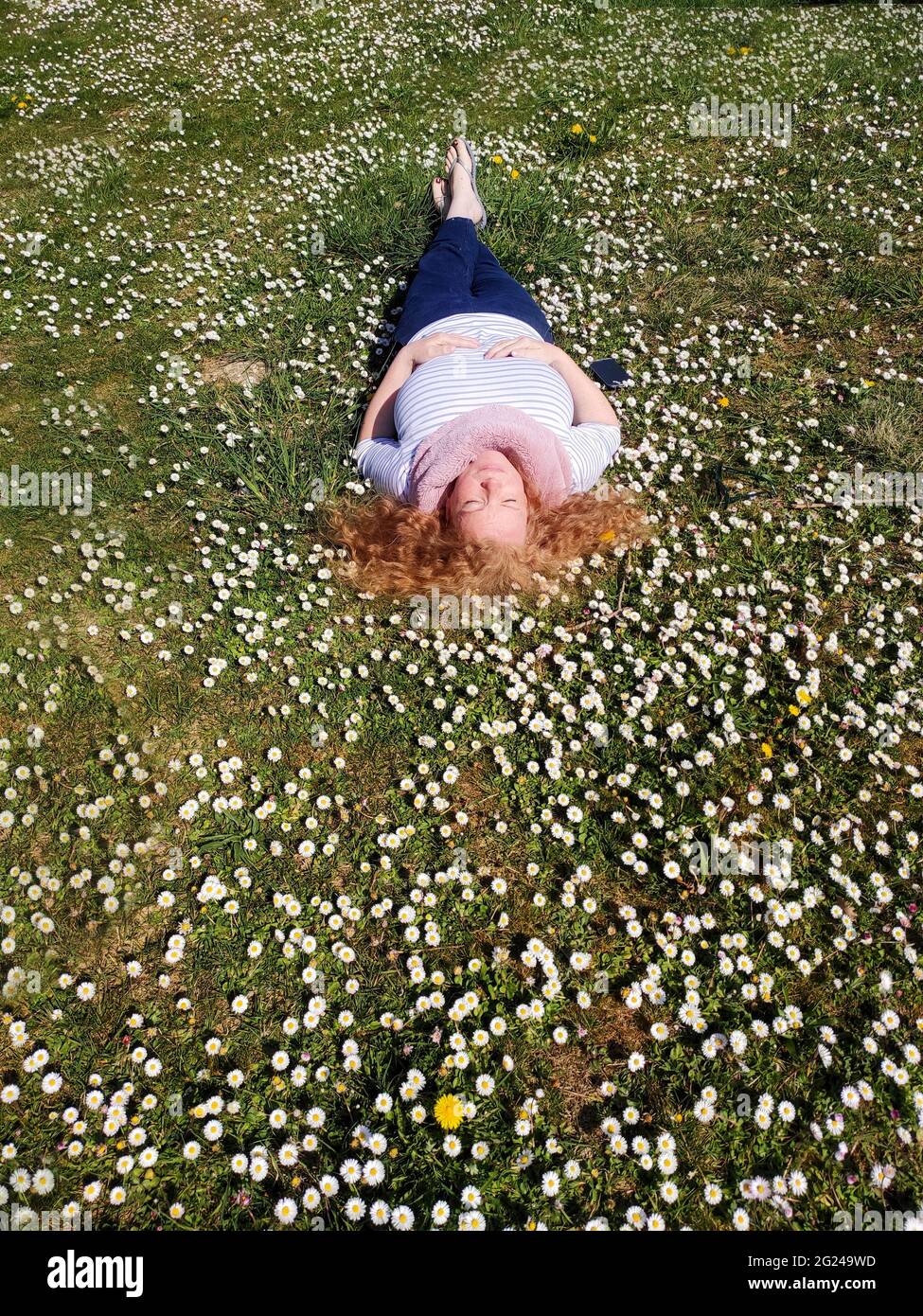 Beautiful redhead mature woman in her fifties lies contentedly in the grass and enjoys the sun and feels good in a field of daisies. Stock Photo