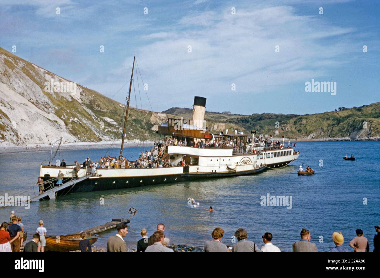 Paddle steamer ‘Consul’ with day trippers arriving from Weymouth disembarking at Lulworth Cove, Dorset, England, UK c. 1958. Lulworth Cove is near the village of West Lulworth, on the Jurassic Coast in southern England. The area is now a World Heritage Site. Consul was built in 1896 and was operated for many years by Cosens of Weymouth. Boats like Consul were designed to run up on the beach and unload passengers via a wooden gangway hinged from the bow. This image is from an old amateur 35mm colour transparency – a vintage 1950s photograph. Stock Photo