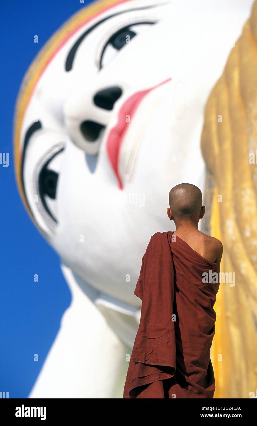 Myanmar, Monyma, Mandalay Division, Novice monk praying under giant statue of reclining Buddha in Lay Kyune Sakkyar temple Stock Photo