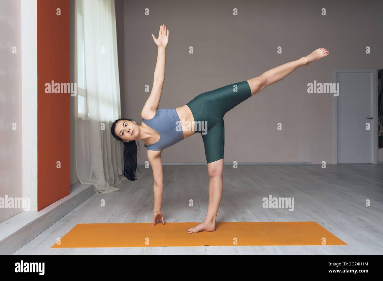 A woman in short leggings and a T-shirt, a yoga practitioner, performs the Ardha Chandrasana exercise, the crescent pose in the studio near the window Stock Photo