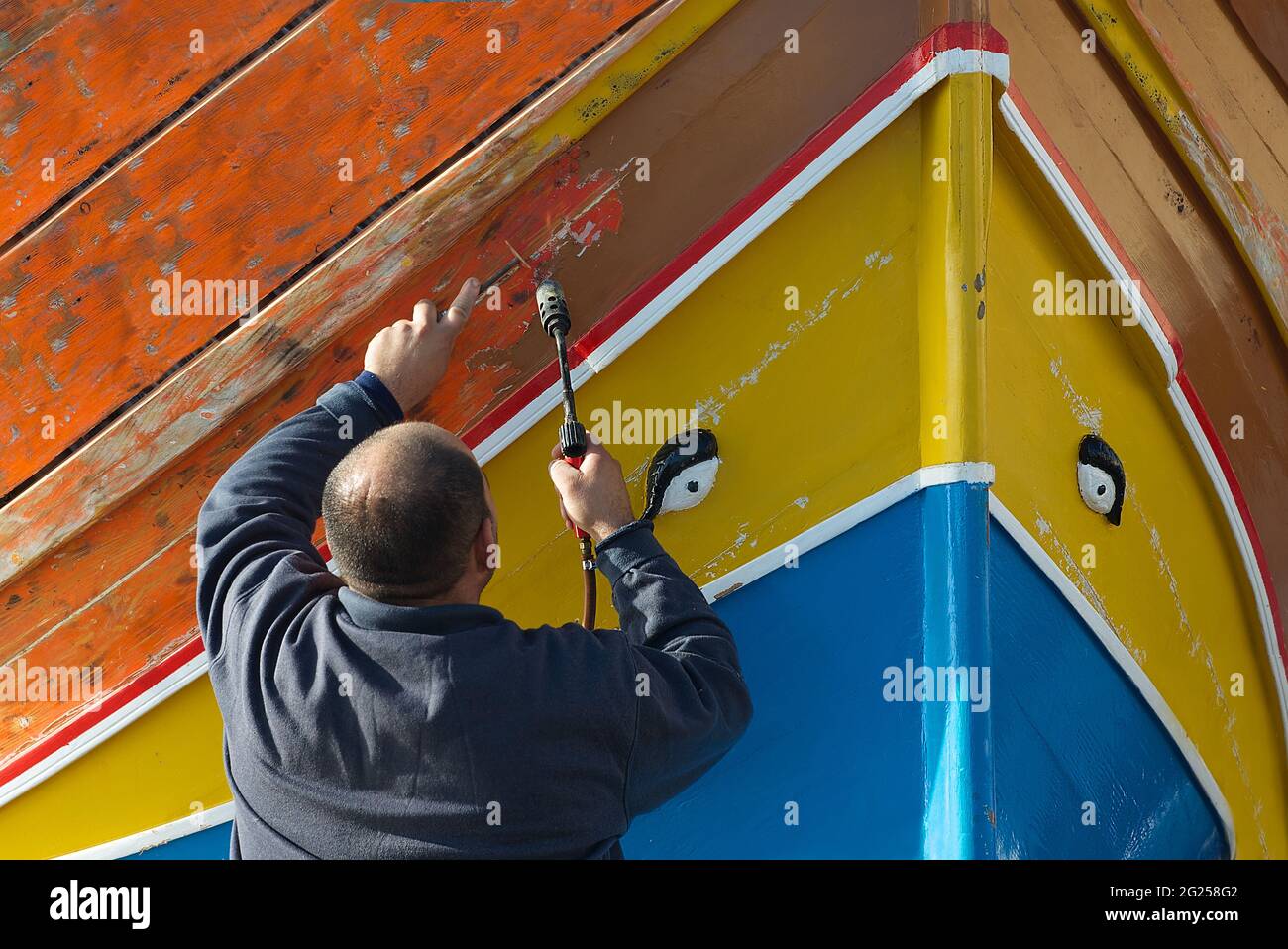 Rear view of a man stripping paint off a traditional luzzu boat with a