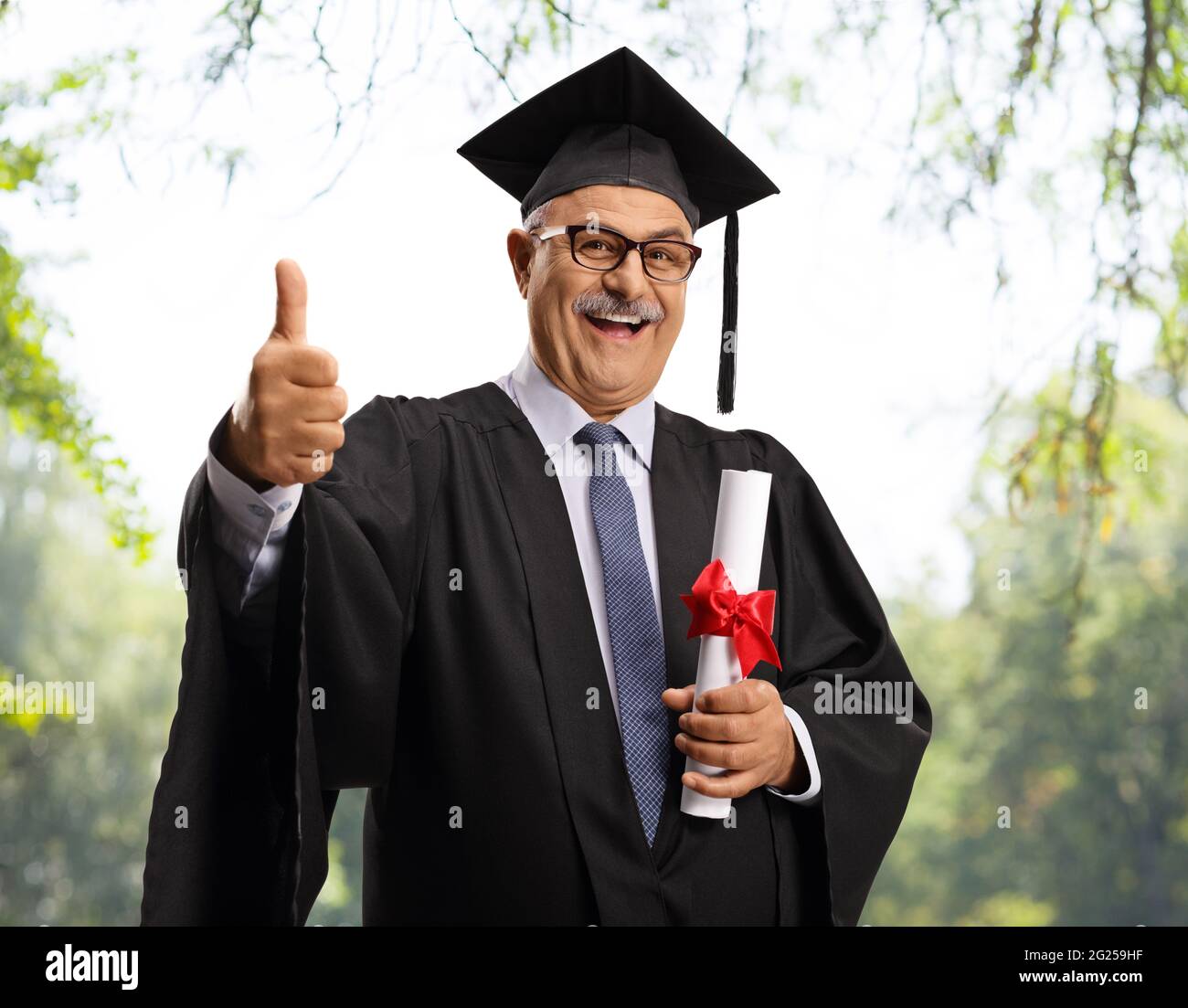 Mature man in a graduation gown holding a diploma and showing thumbs up outdoors Stock Photo