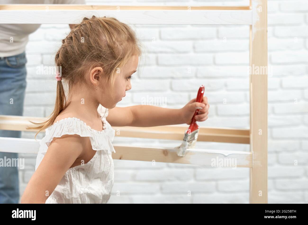 Pretty little child with blond hair painting wooden shelves with brush and white color. Cute kid helping mother to restoring furniture at home. Stock Photo