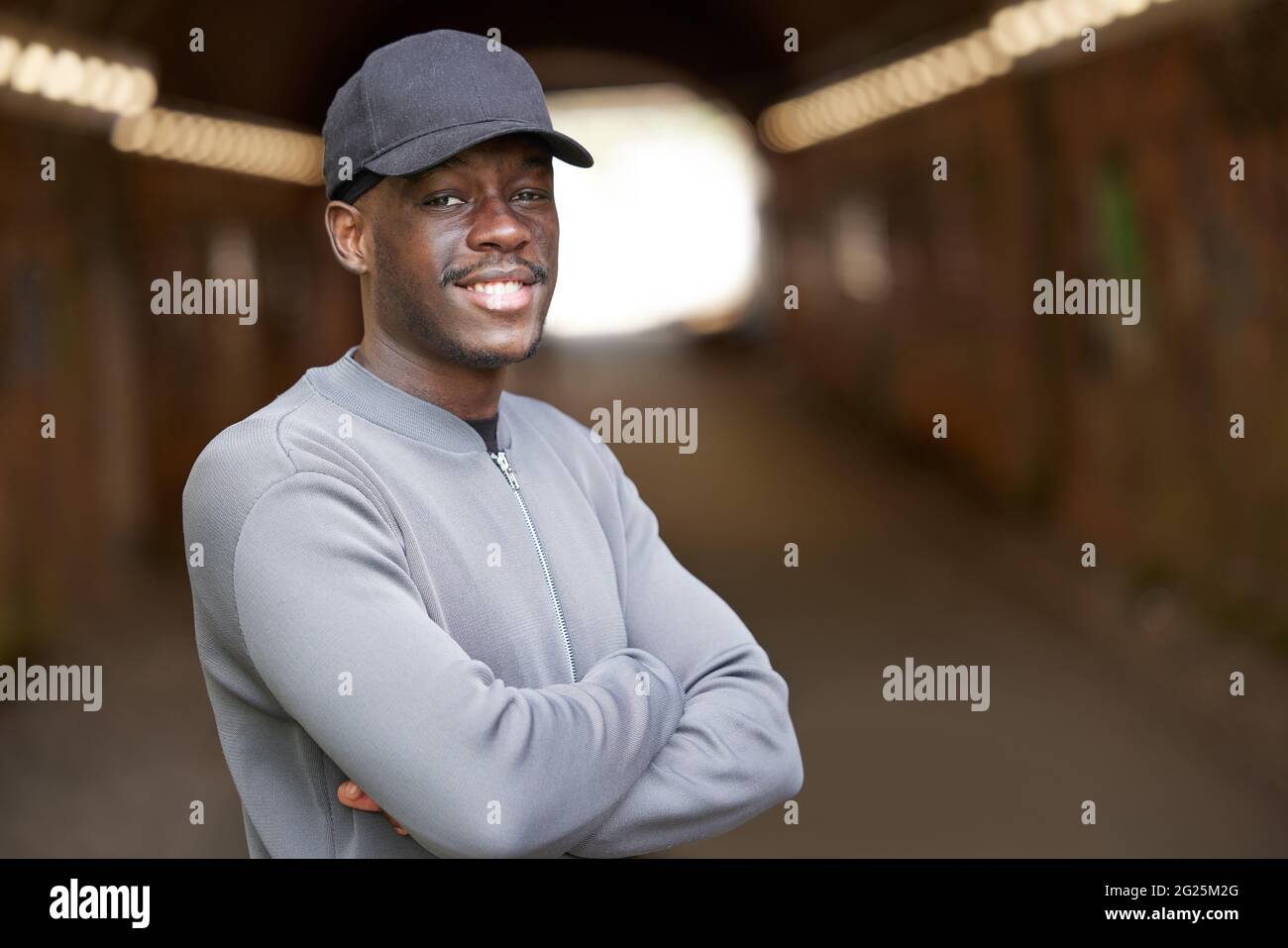 Portrait Of Smiling Young Man Wearing Baseball Cap Standing In Tunnel In In Urban Setting Stock Photo