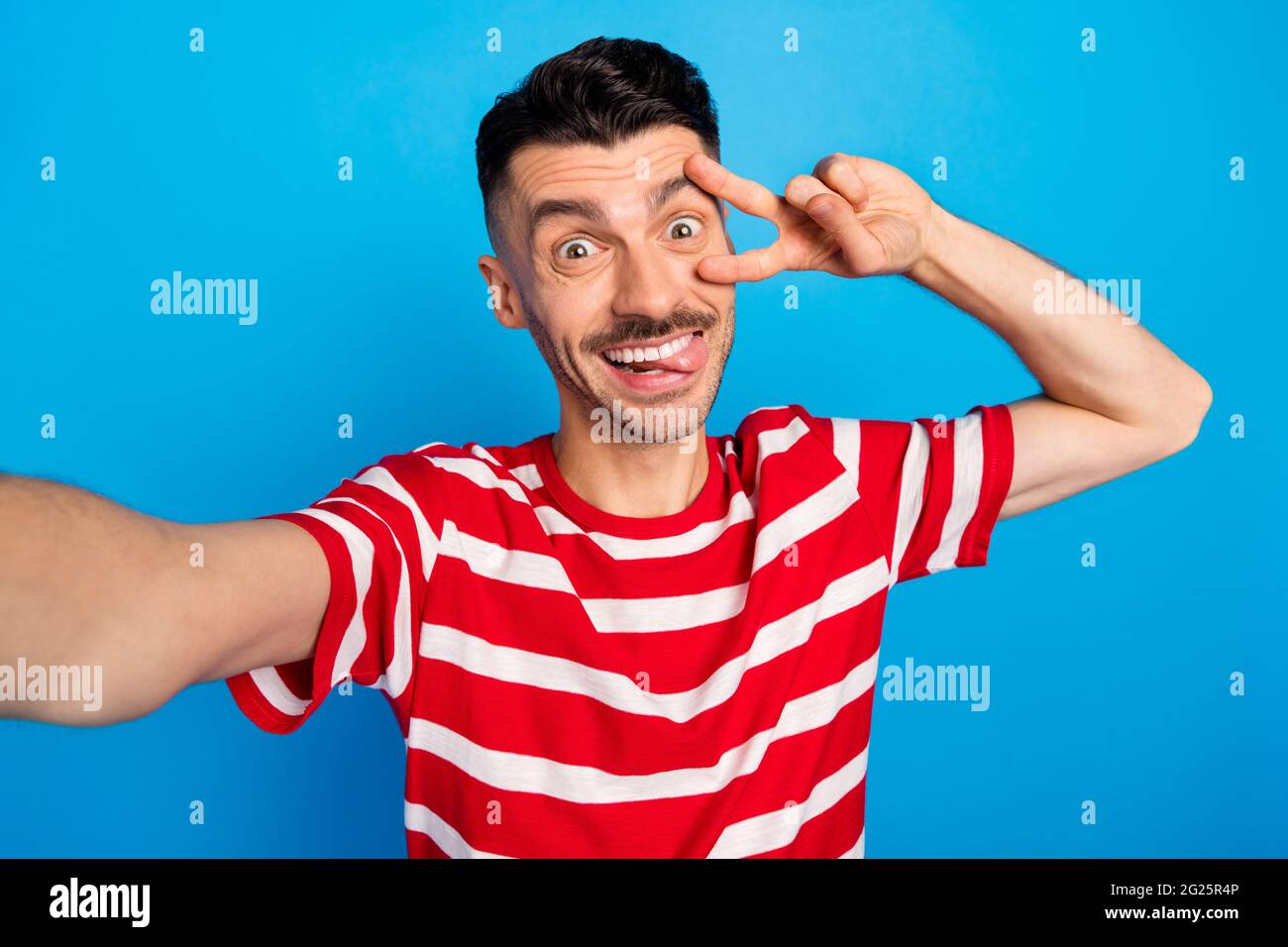 Photo of charming positive young man make v-sign bite tongue make selfie  isolated on blue color background Stock Photo - Alamy