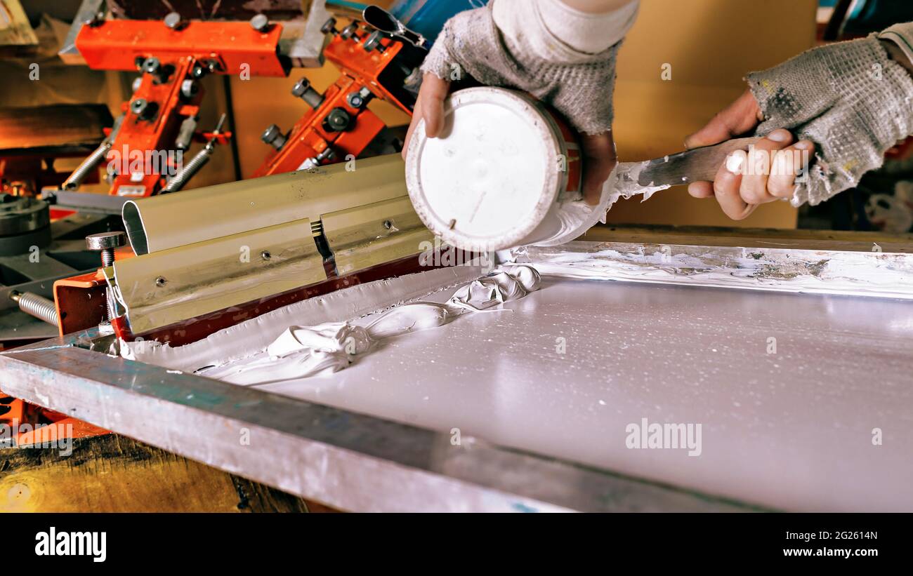 selective focus photo of print screening apparatus, can of white paint and hands of a worker. serigraphy production. printing images on clothes by sil Stock Photo
