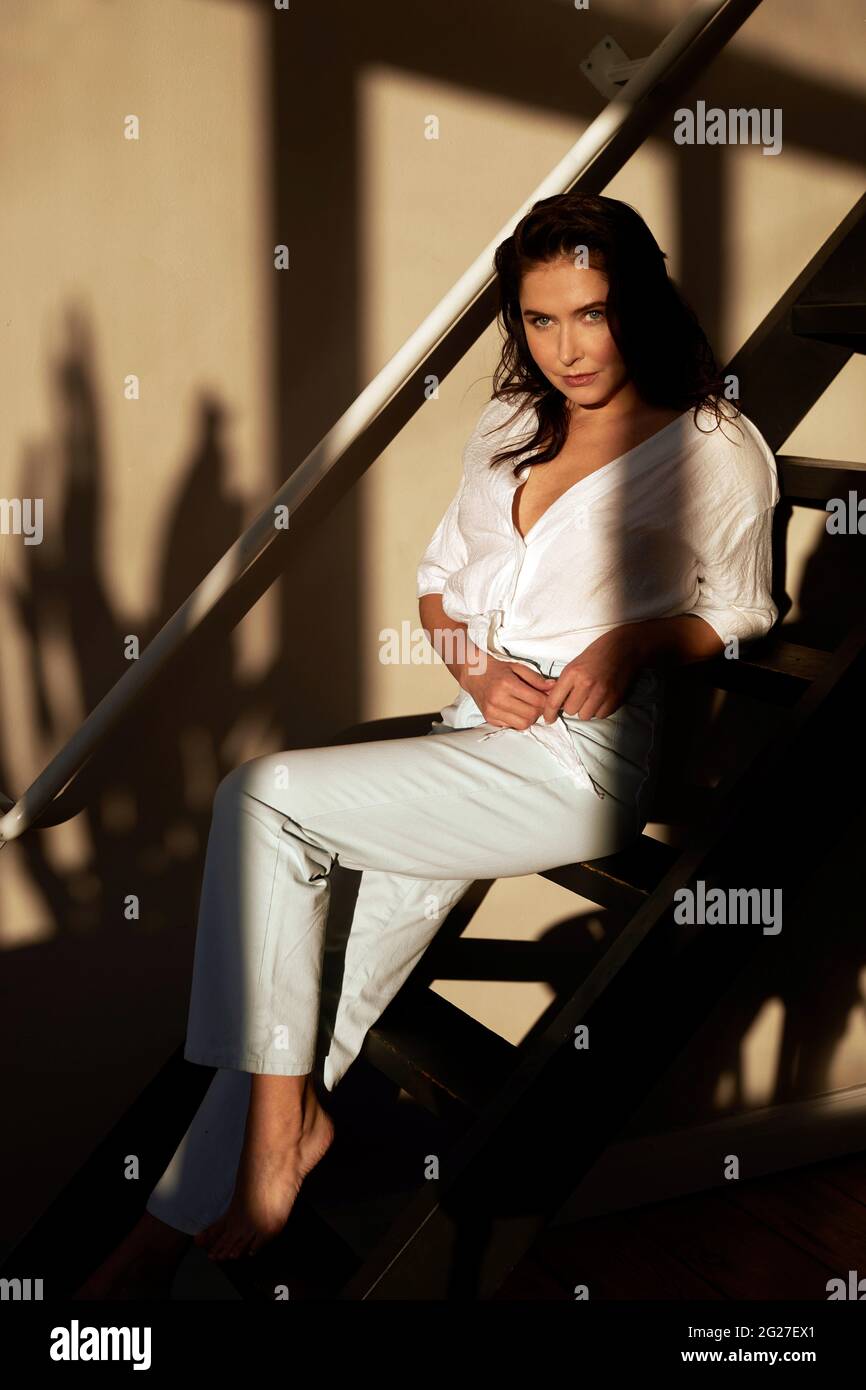 Young confident female wearing white shirt and pants sitting on wooden stairway and looking at camera Stock Photo