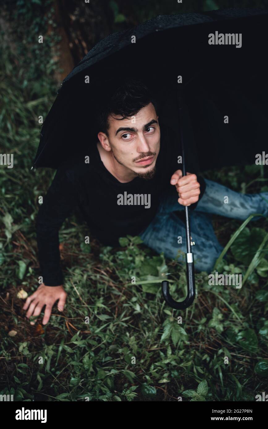 Young handsome man in the forest holding a black umbrella and lying on the grass Stock Photo