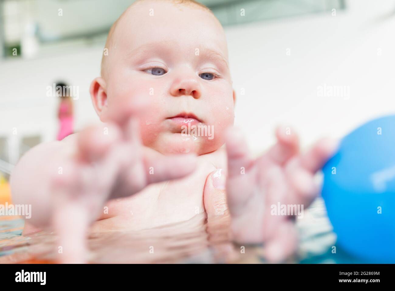 Newborn baby in pool at infant swimming lesson reaching for water ball
