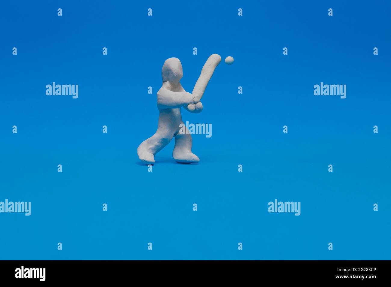 A white plasticine doll practicing baseball on a blue background. The doll is a batter and holds the bat ready to hit the ball coming towards him. Stock Photo