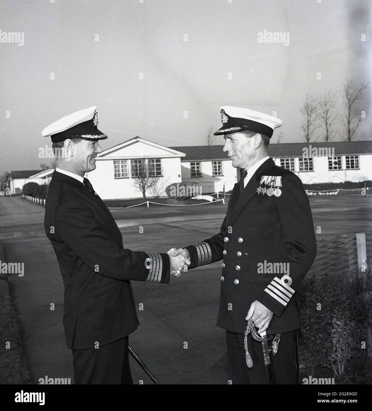 1960s, historical, outside at HMS Calendonia, two Royal Naval  Captains shaking hands, in a hand-over of command, Fife, Scotland. Captain (Capt) is a senior officer rank of the Royal Navy, above a Commander and below Commodore and has a NATO ranking code of OF-5. The rank is equivalent to a Colonel in the British Army and Royal Marines, and to a Group Captain in the Royal Air Force. HMS Caledonia was first opened in 1937 and was responsible for artificer apprentice training up to 1985, with many thousands of young men going through training. Stock Photo