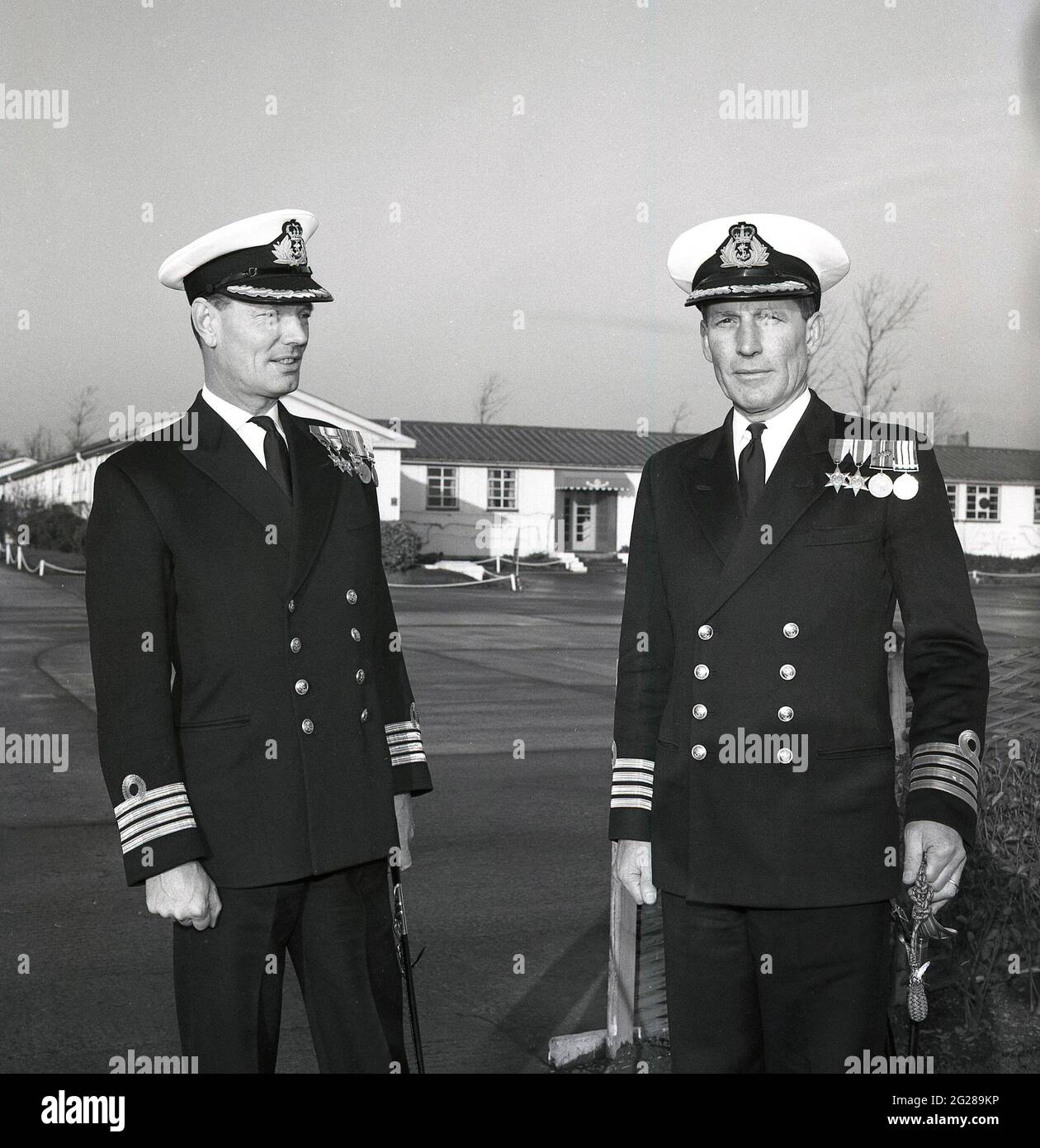 1960s, historical, outside HMS Caledonia, two decorated Royal Naval Captains in full uniform pose for a photograph in a hand-over of command, Fife, Scotland. Captain (Capt) is a senior officer rank of the Royal Navy, above a Commander and below Commodore and has a NATO ranking code of OF-5. The rank is equivalent to a Colonel in the British Army and Royal Marines, and to a Group Captain in the Royal Air Force. HMS Caledonia was first opened in 1937 and was responsible for artificer apprentice training up to 1985, with many thousands of young men going through training. Stock Photo
