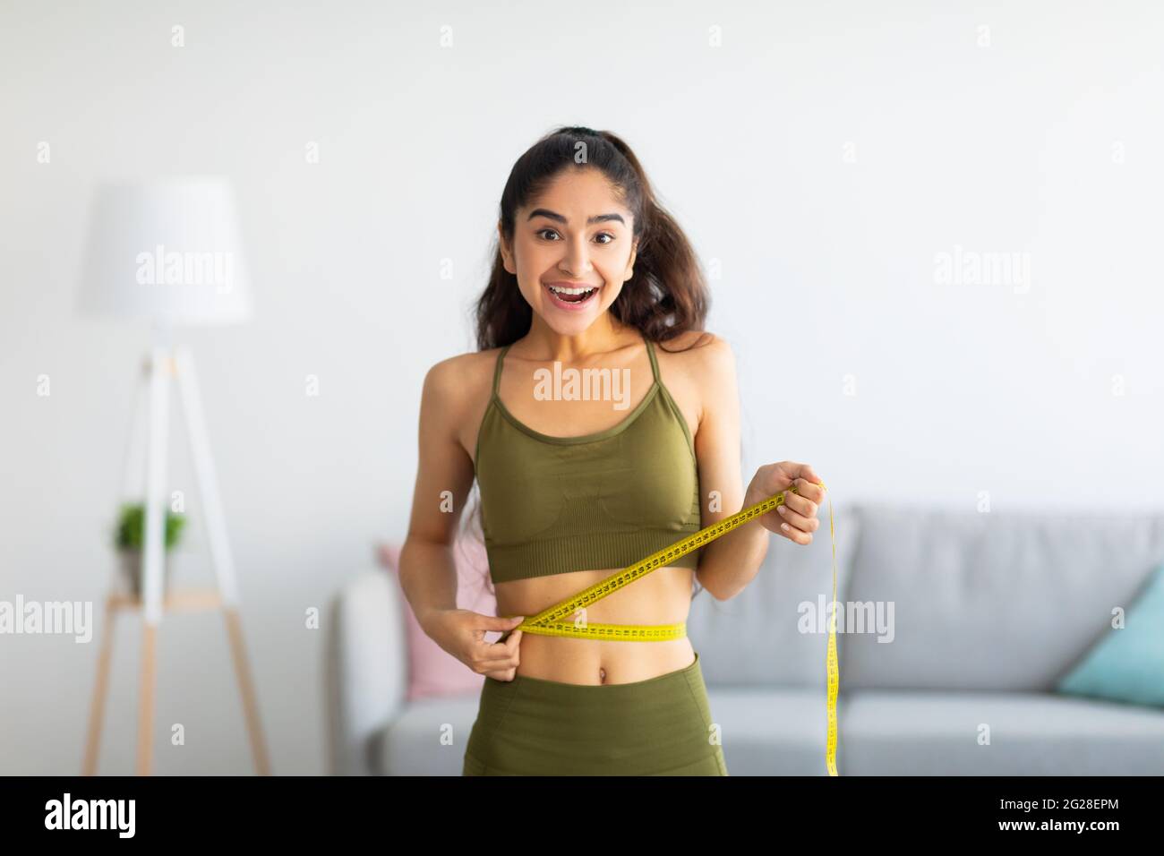 Portrait of young Indian woman measuring her waist with tape indoors, smiling at camera Stock Photo