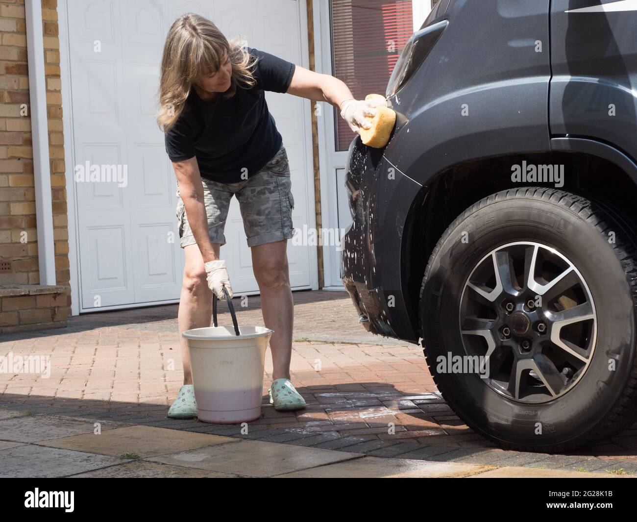 A lady motorhome owner holds bucket handle and cleans her recreational vehicle with a sponge.She wears shorts on a sunny day - Stock Photo