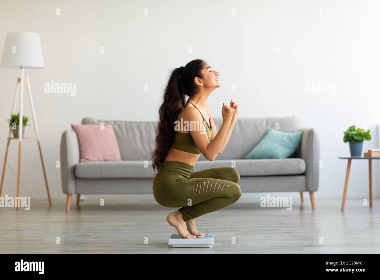 Full length of excited Indian woman sitting on scales at home, overjoyed with success of her slimming diet, side view Stock Photo
