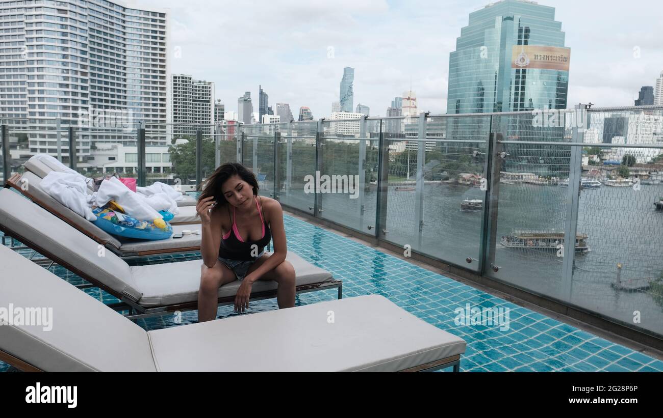 Young Lady Dark Hair wearing Black Top and Light Blue Denim cut-off Shorts Sitting on Deck Chair at Roof Top Swimming Pool Bangkok Thailand Stock Photo