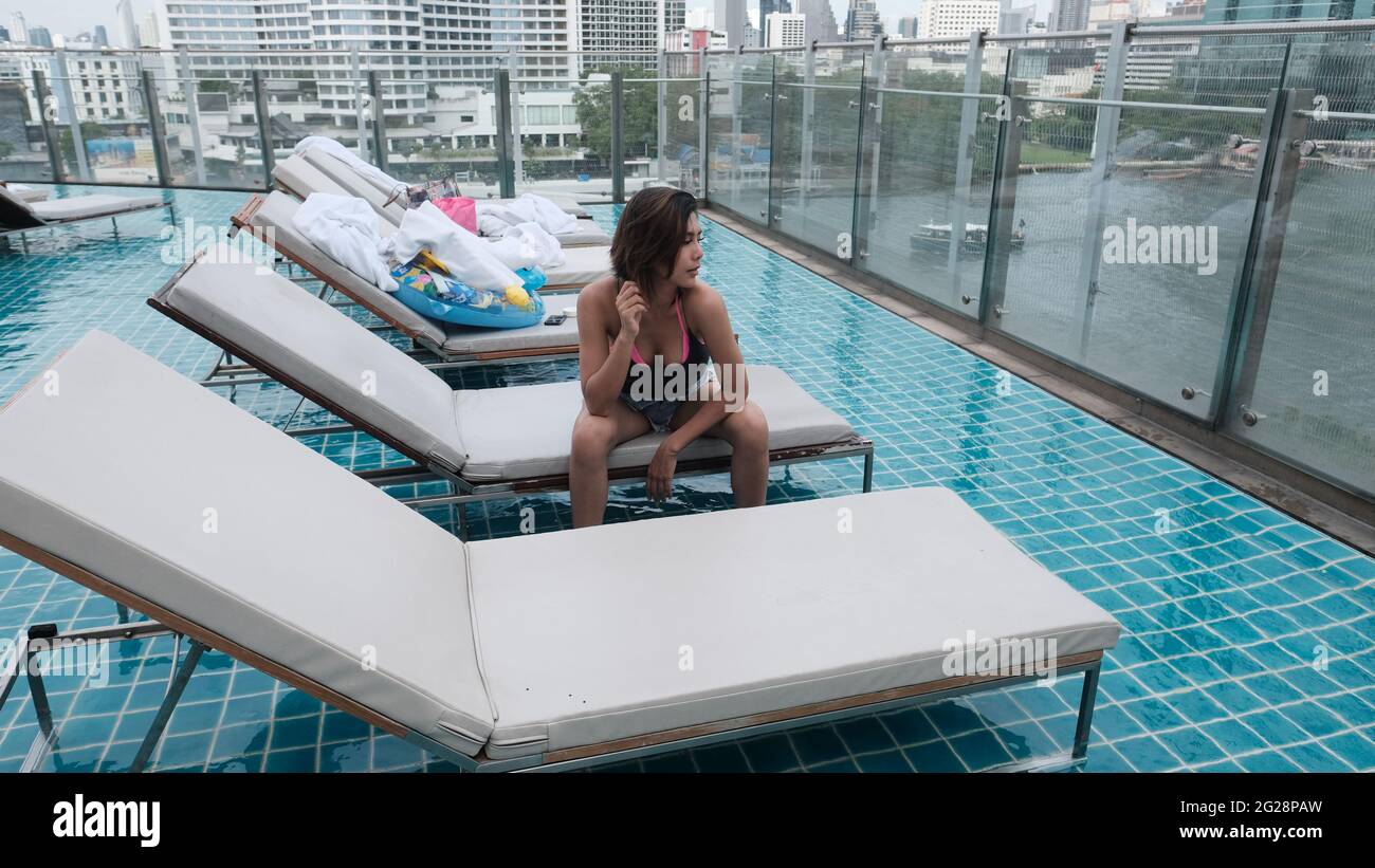 Young Lady Dark Hair wearing Black Top and Light Blue Denim cut-off Shorts Sitting on Deck Chair at Roof Top Swimming Pool Bangkok Thailand Stock Photo