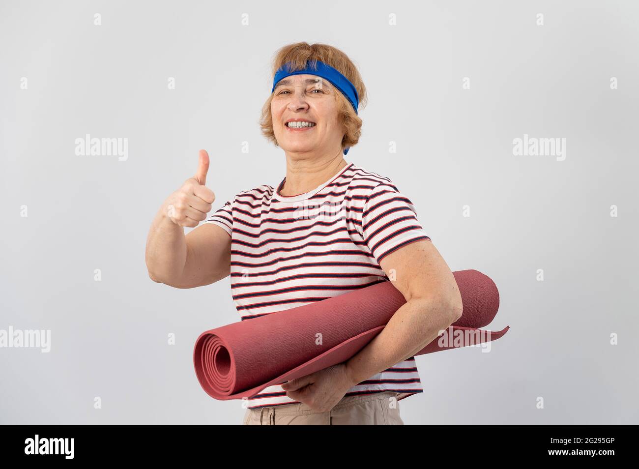 An elderly woman holds a yoga mat and shows her thumb up. The pensioner approves of sports. Stock Photo