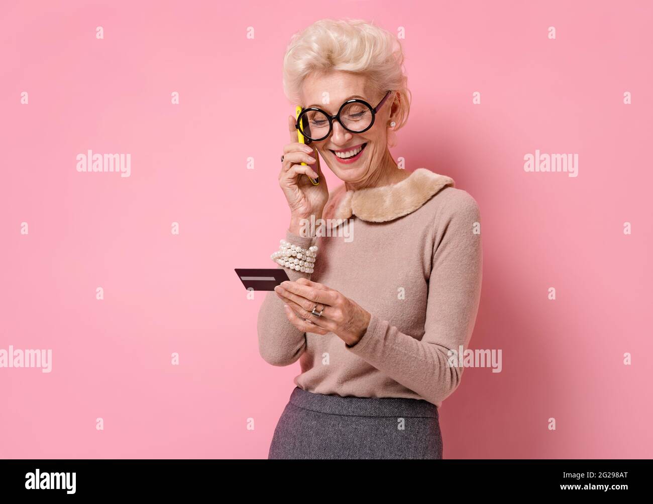 Woman reading something off her credit card while talking on the phone. Photo of kind elderly woman on pink background Stock Photo