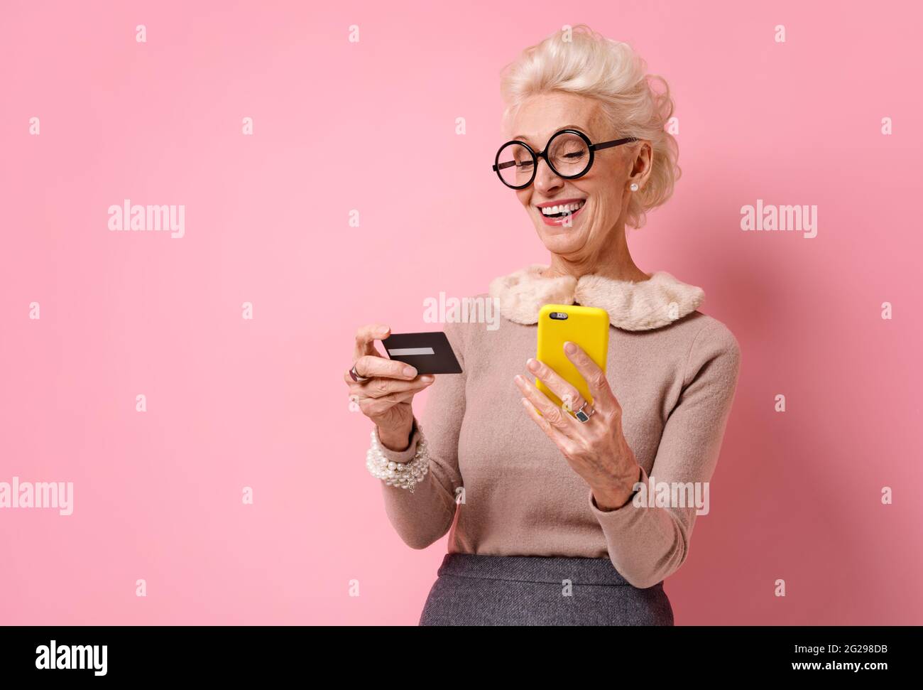 Grandmother makes a payment, using a credit card and smartphone. Photo of kind elderly woman on pink background. Stock Photo
