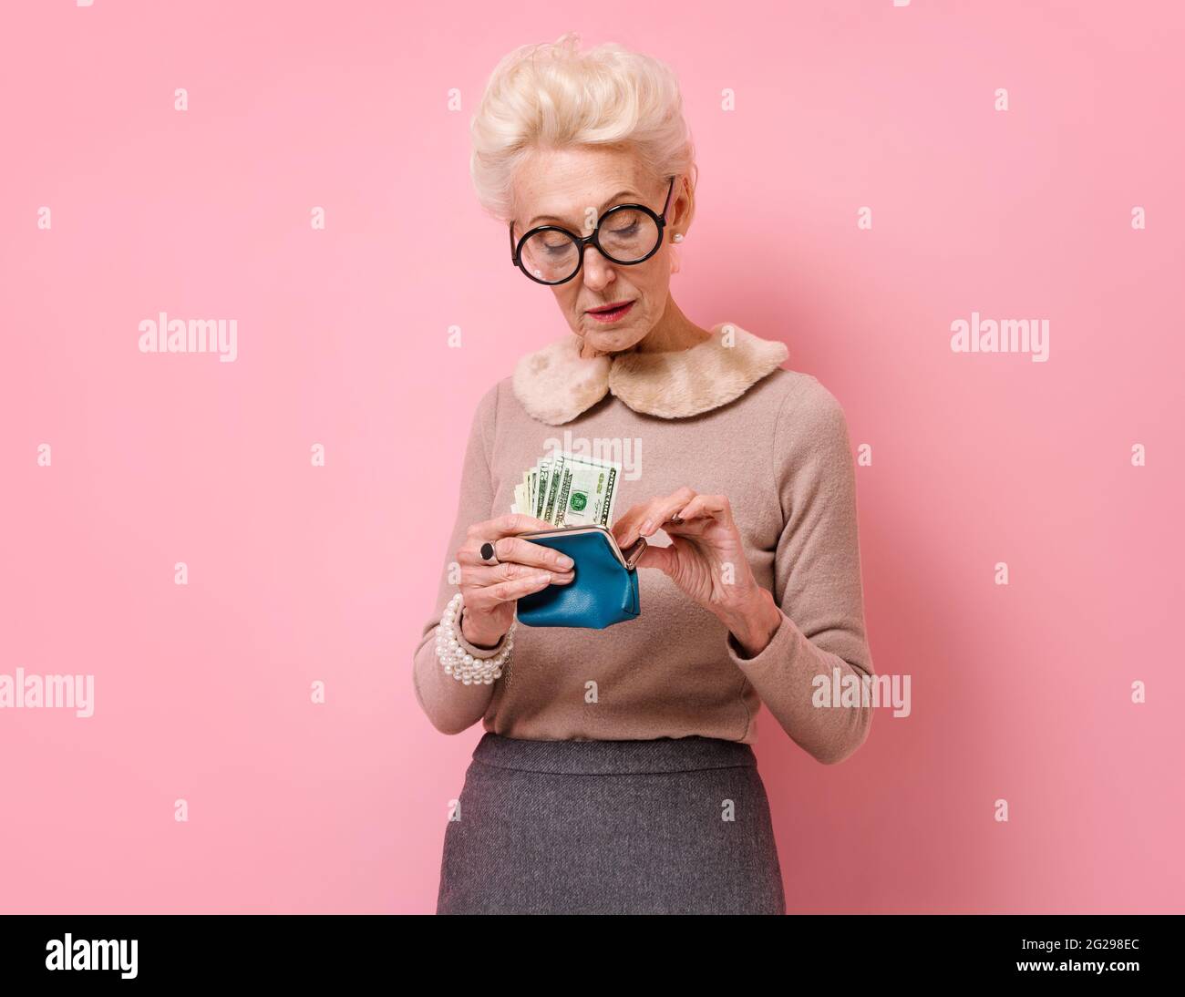 Sad woman looking at her wallet. Photo of kind elderly woman wears eyeglasses on pink background. Stock Photo