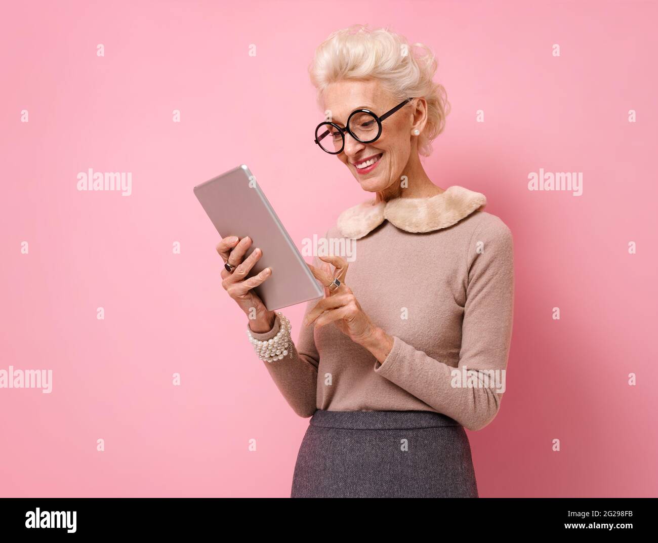 Grandmother uses the tablet, checks for news. Photo of kind elderly woman on pink background. Stock Photo