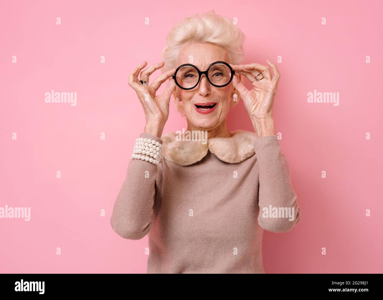 Overjoyed grandmother laughs at something funny. Photo of kind elderly woman in eyeglasses looking at camera on pink background. Stock Photo