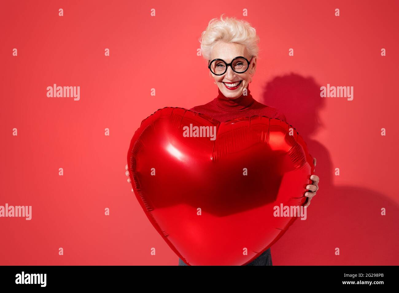 Happy woman holds red heart shape balloon. Photo of smiling elderly woman in love on red background. Valentine's Day Stock Photo