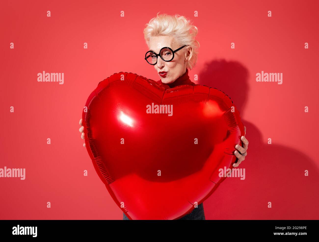 Happy woman holds red heart shape balloon. Photo of smiling elderly woman in love on red background. Valentine's Day Stock Photo