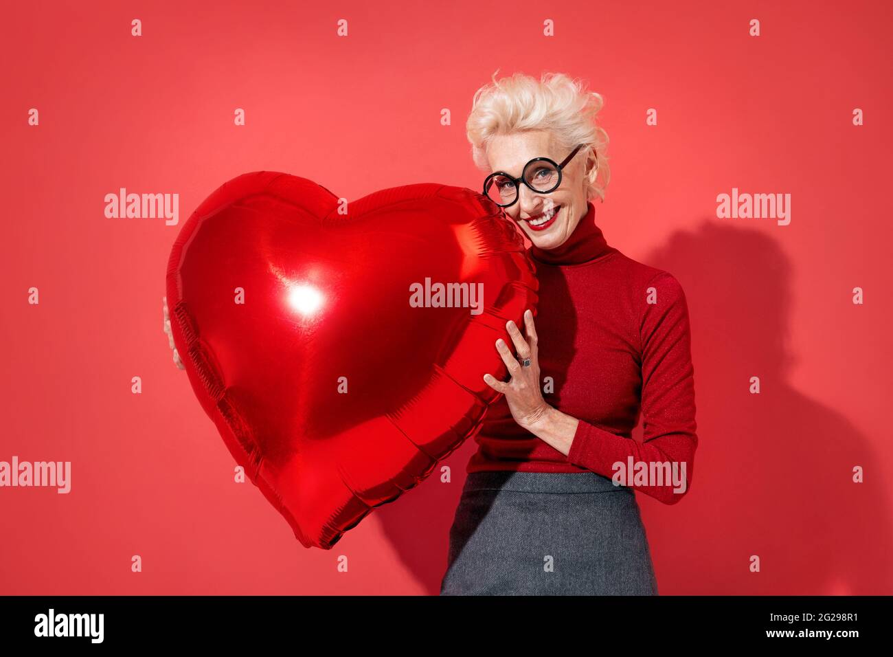 Lovely woman hugs red heart shape balloon. Photo of smiling elderly woman in love on red background. Valentine's Day Stock Photo