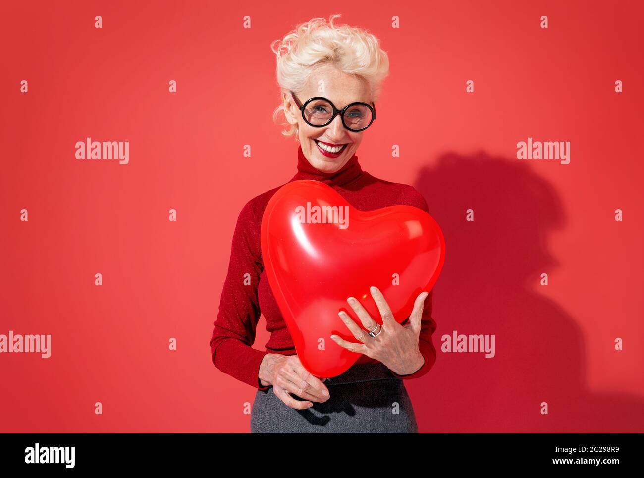 Lovely woman hugs red heart shape balloon. Photo of smiling elderly woman in love on red background. Valentine's Day Stock Photo