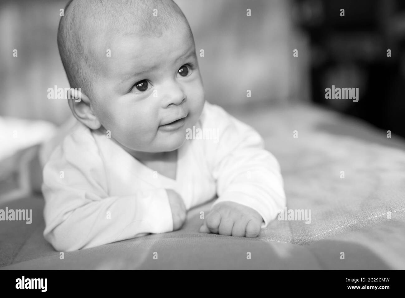 Adorable baby in sunny bedroom. Newborn baby is resting in bed covered warm soft blanket. Smiling child looking at camera. Stock Photo