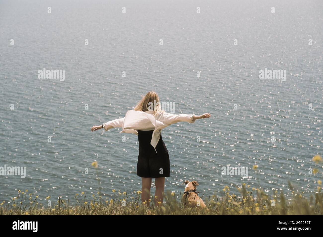 Woman next to a dog enjoying the sea scenery, arms spread. Going Stock Photo