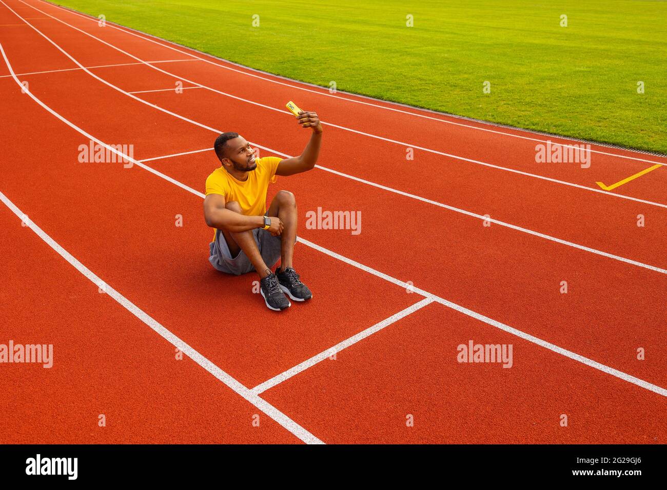 Handsome young afro american sportsman athlete runner is taking selfie at the stadium location. Smiling joyful black male laying on the field and Stock Photo