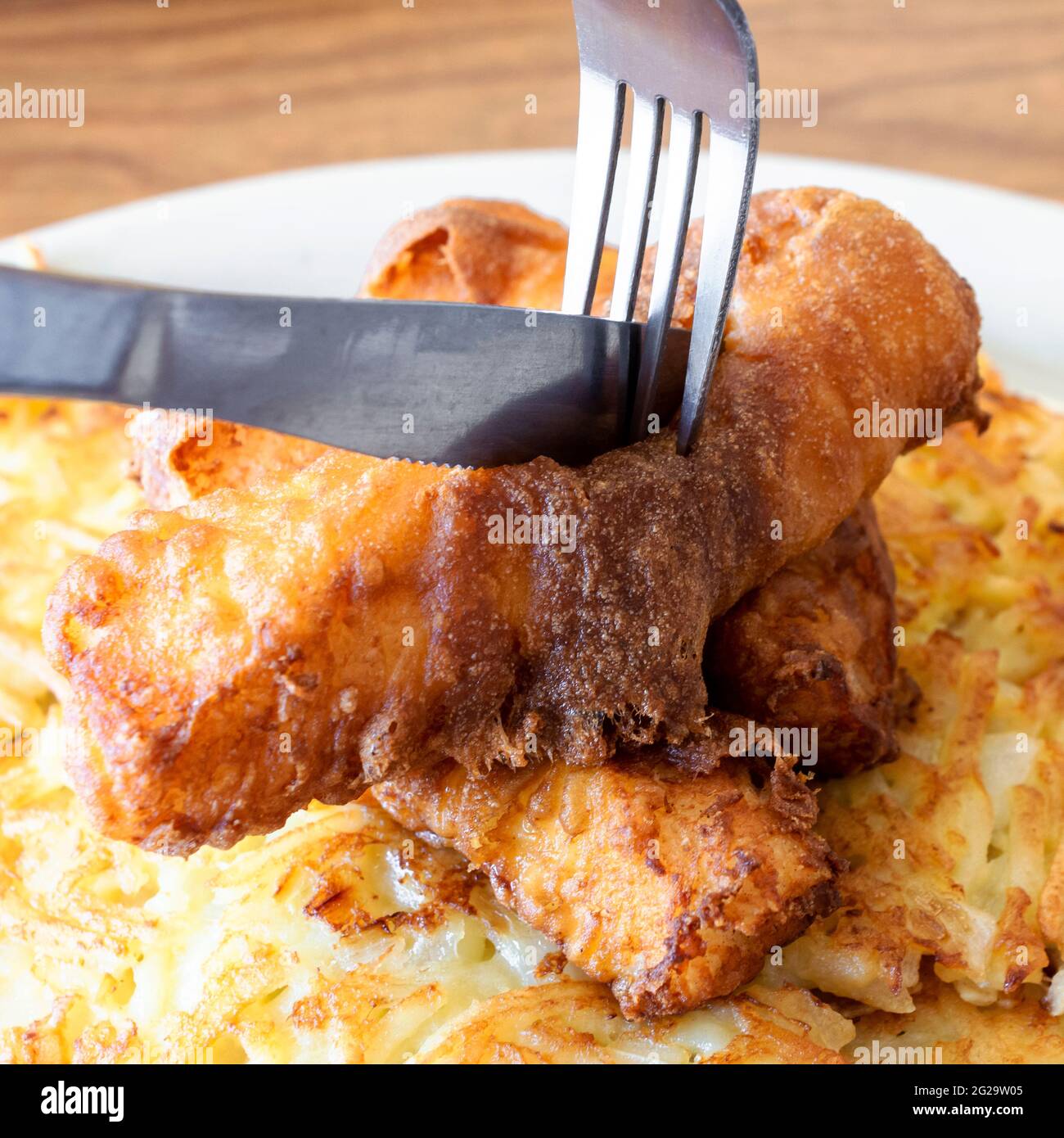 Deep fried fish is being cut into while on a bed of hashbrowns on a white plate. Stock Photo