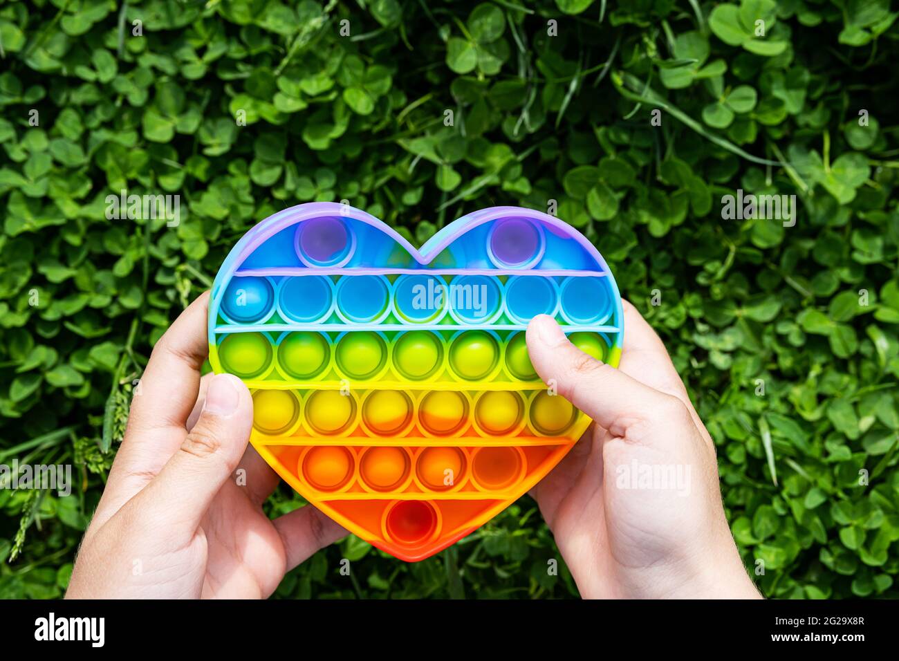 Child plays on the grass with a simple dimple relaxing  toy. Pop it toy heart shaped, rainbow colored in boys hands at summer day. Stock Photo