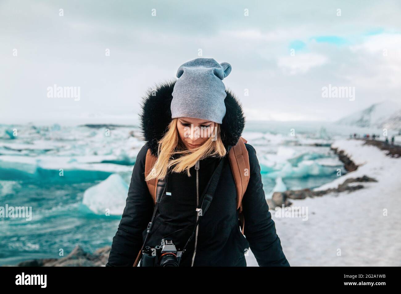 Blond haired Woman in hat and warm jacket looking down standing on shore of frozen turquoise sea and snowy coast in Iceland Stock Photo