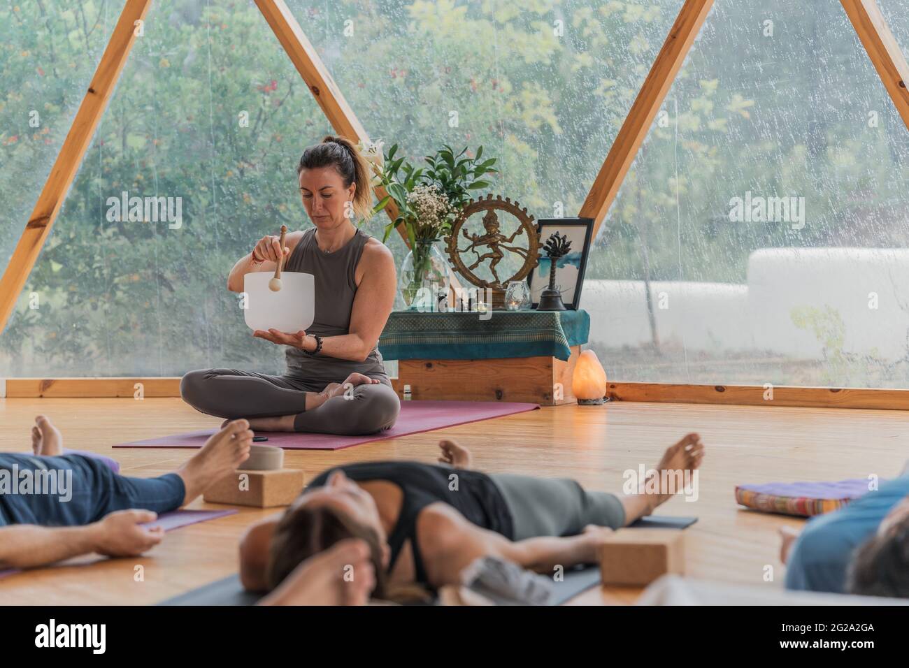Relaxed people lie in shavasana with closed eyes on yoga mat while concentrated coach extracting sound from singing bowl in round class for yoga Stock Photo