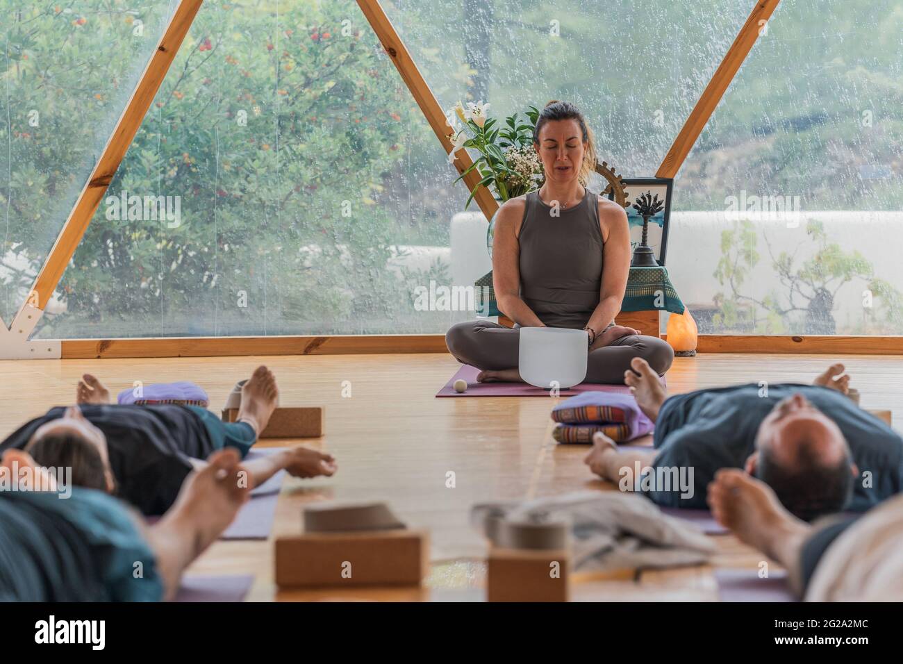 Relaxed people lie in shavasana with closed eyes on yoga mat while concentrated coach extracting sound from singing bowl in round class for yoga Stock Photo