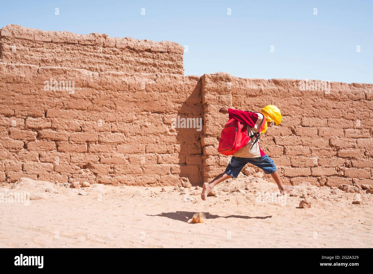 Side view of anonymous barefoot black boy with backpack running to school near shabby brick wall on sunny day in Sahara Desert Stock Photo