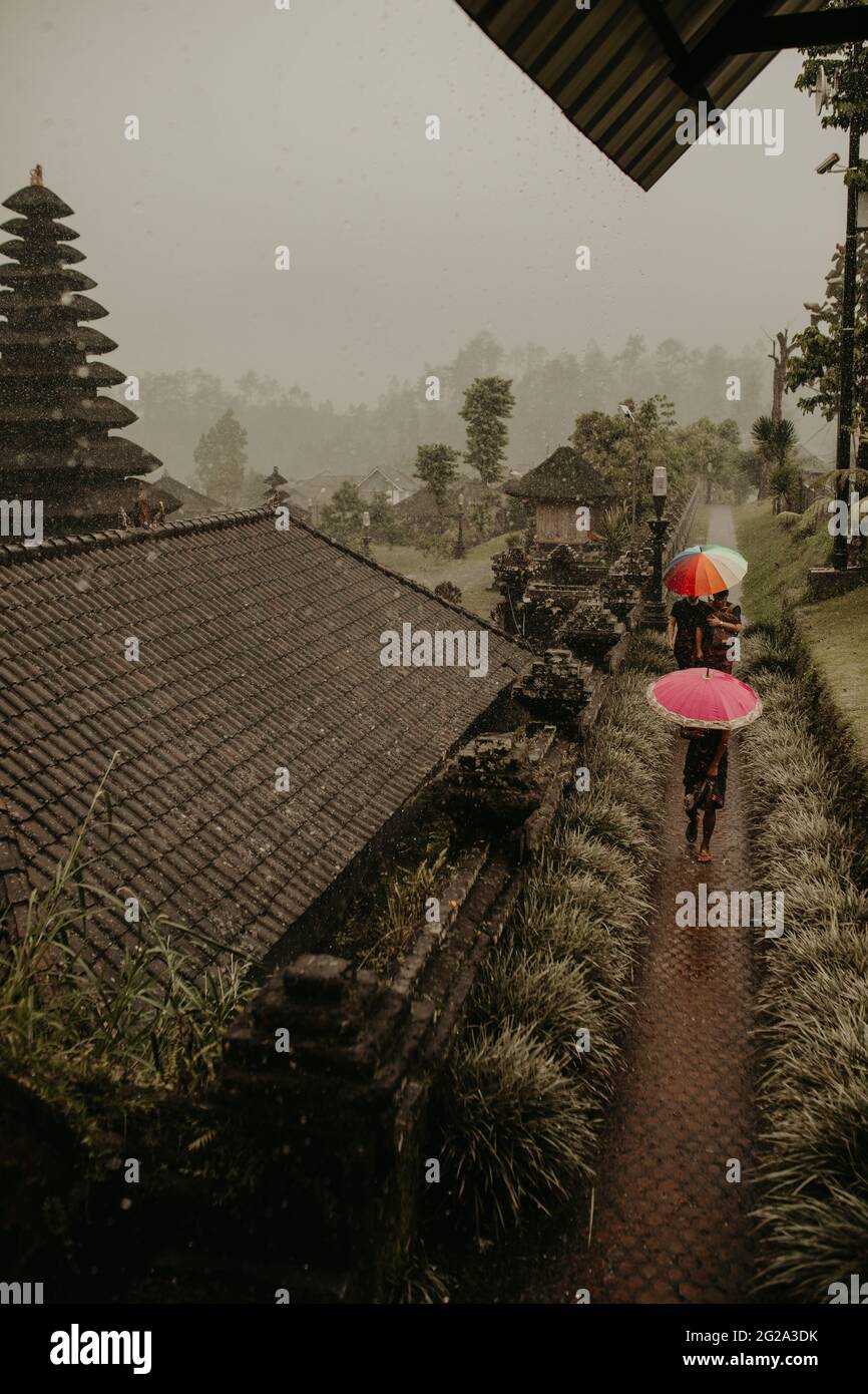 Unrecognizable people with umbrellas walking on wet path near Asian houses on rainy day in traditional village in Bali Stock Photo