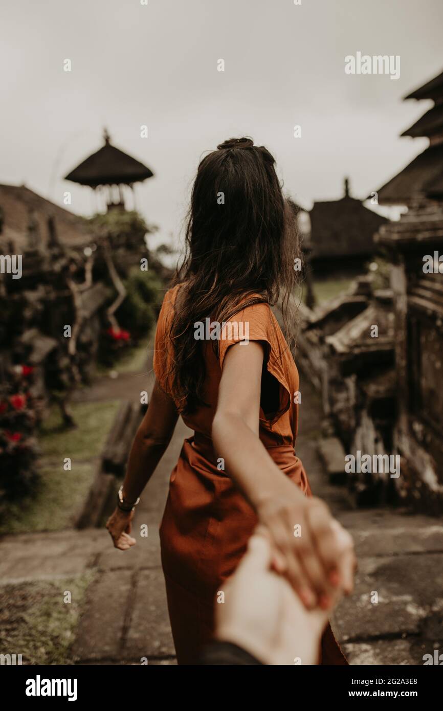 Back view of female holding crop hand of friend and asking to follow her on old street of oriental town In Bali Stock Photo