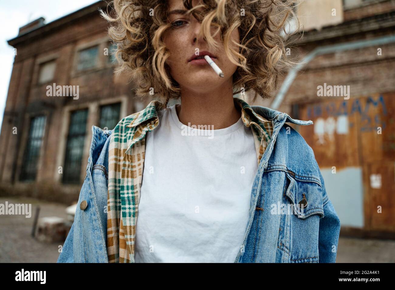 Stylish curly Woman standing and thoughtfully smoking in abandoned place looking at camera Stock Photo