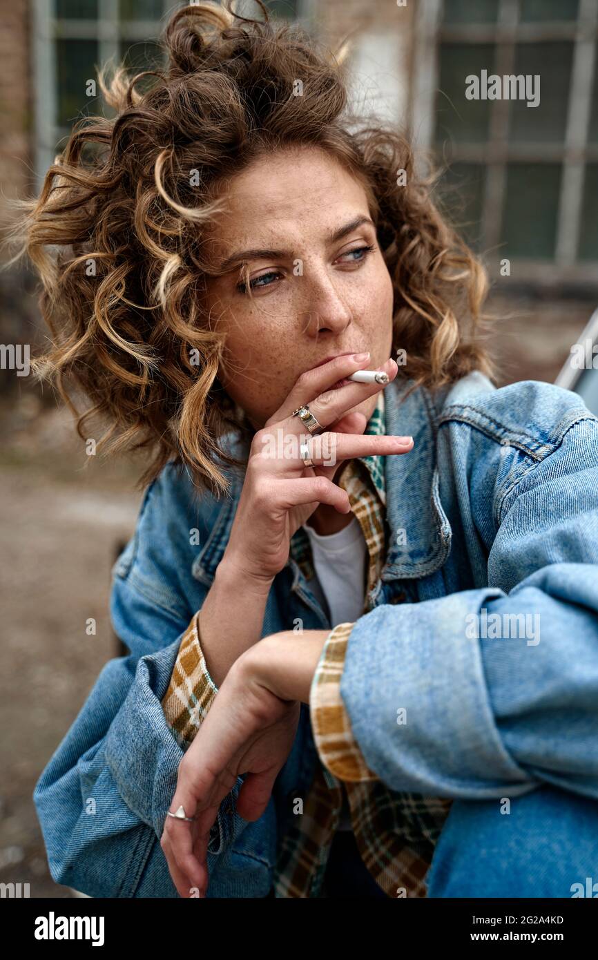 Stylish curly Woman sitting and thoughtfully smoking in abandoned place looking along Stock Photo