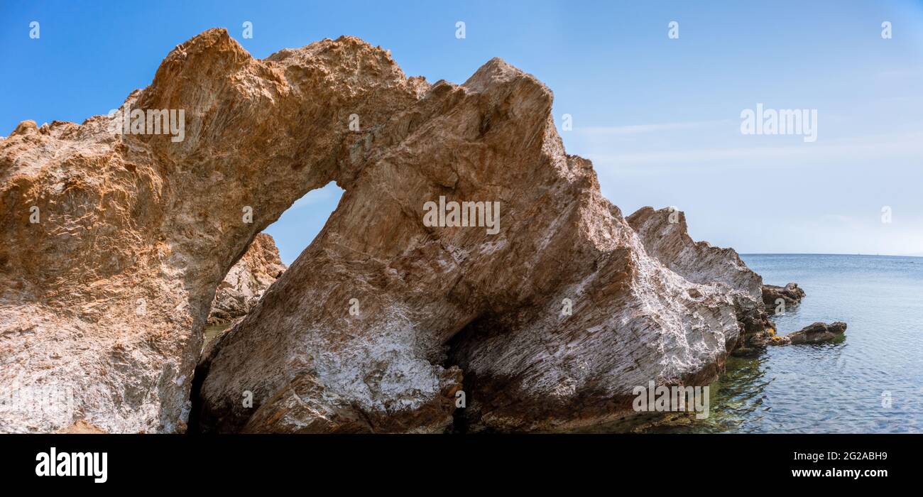 Big rock with a hole on wild mediterranean sea shore with crystal clear water and blue sky. Travel Greece near Athens. Summer nature scenic lagoon pan Stock Photo