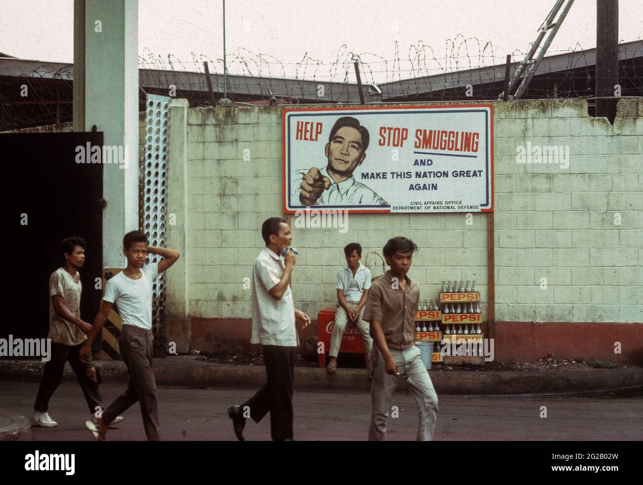 Manila, 1966. Filipino dock workers stroll past a billboard appeal from President Ferdinand Marcos to 'Help Stop Smuggling and Make This Nation Great Again''. Contraband goods were being openly handled nearby. Stock Photo