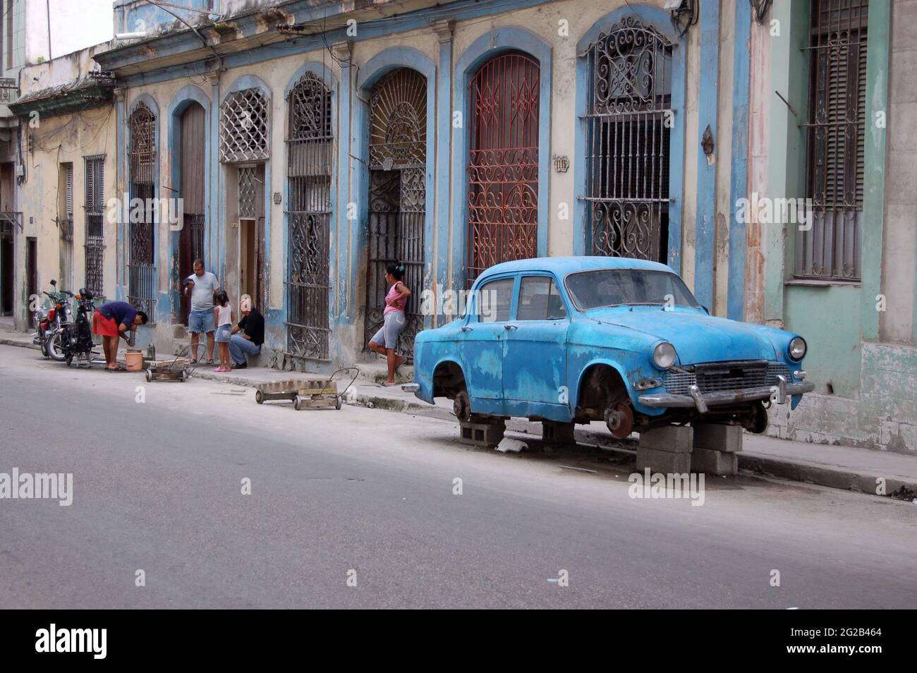 Havana, Cuba - November 19, 2005:  Street scene in Central Havana as a mechanic repairs a motorbike in the road with a battered vintage car resting on Stock Photo